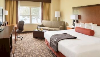 A cozy hotel room with a large bed, a sofa, a desk with a chair, and a window with a view. A TV is mounted in front of the bed, ending the sentence.