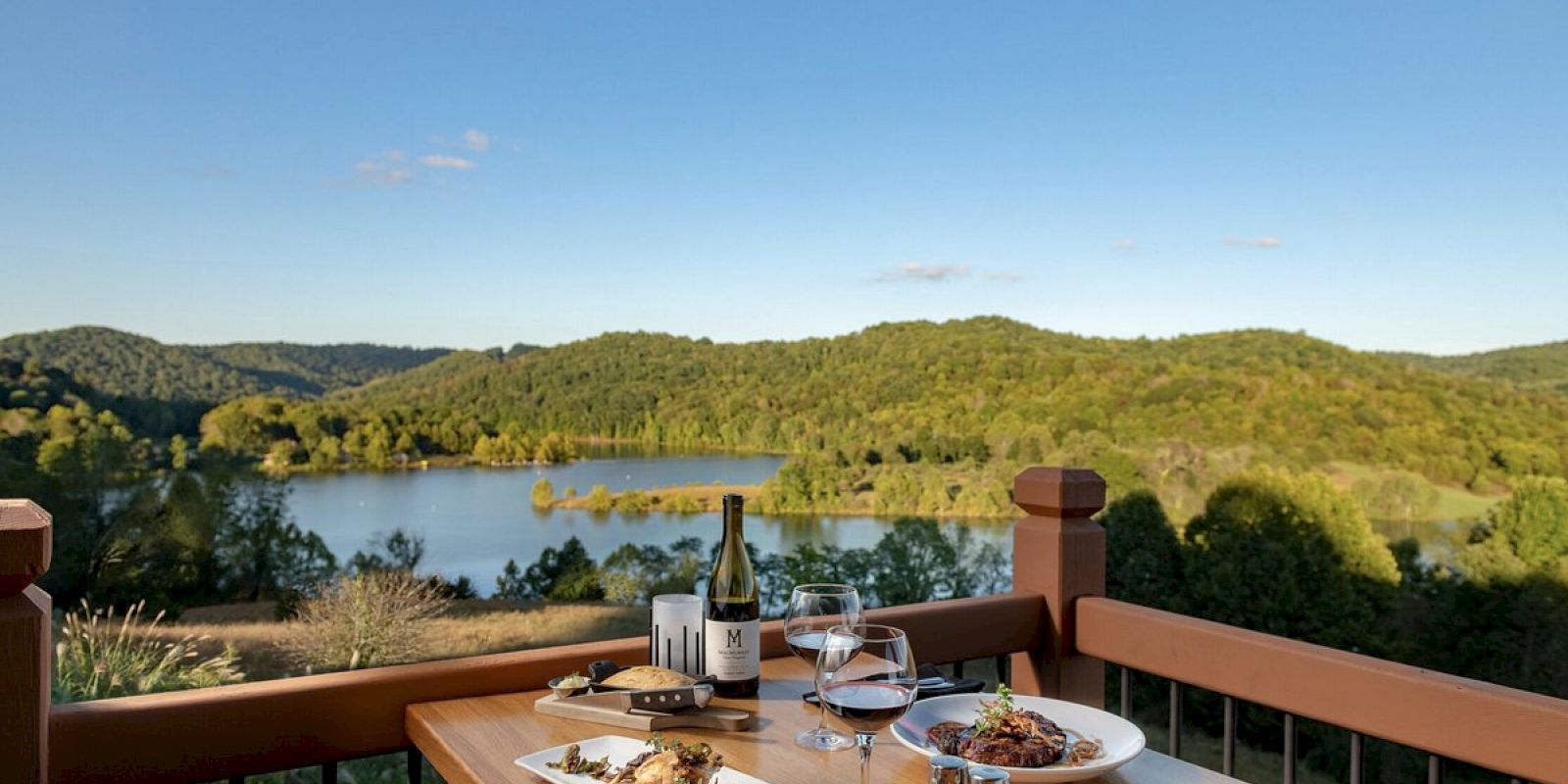 A wooden table on a deck with plates of food, wine glasses, and a bottle, overlooking a lake and forested hills under a clear blue sky.