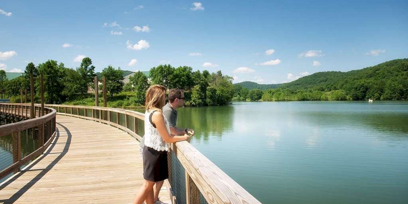 Two people stand on a wooden boardwalk overlooking a calm lake surrounded by green hills and trees, under a clear blue sky.