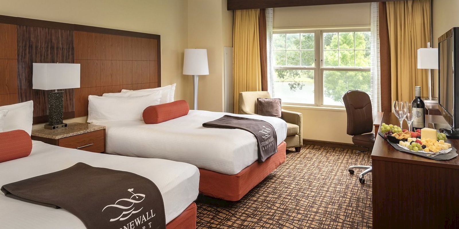 The image shows a neatly arranged hotel room with two beds, a flat-screen TV, an armchair, a window with curtains, and a snack tray.