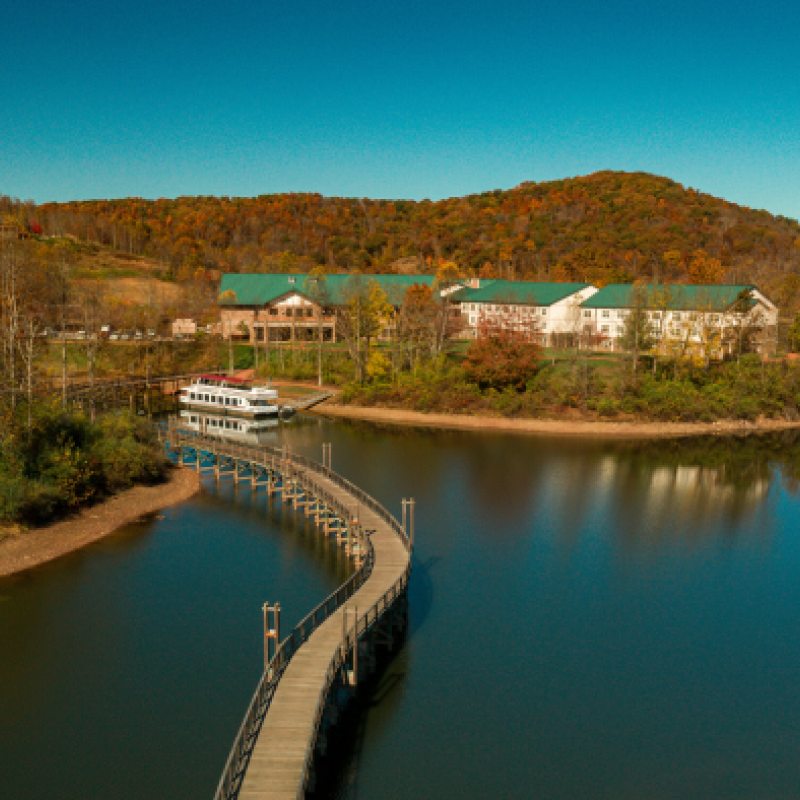 The image shows a serene lake with a curved wooden walking bridge, surrounded by autumnal trees and hills, leading to buildings with green roofs.