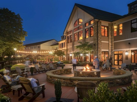 The image shows a cozy outdoor seating area at dusk with a fire pit, surrounded by Adirondack chairs, string lights, and a large, well-lit building behind.