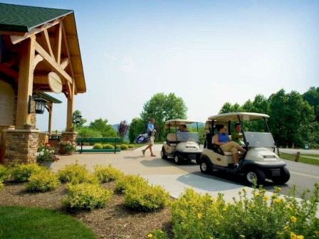 A paved area with two golf carts and people around them, set near a building with a wooden structure and surrounded by green plants and shrubs.