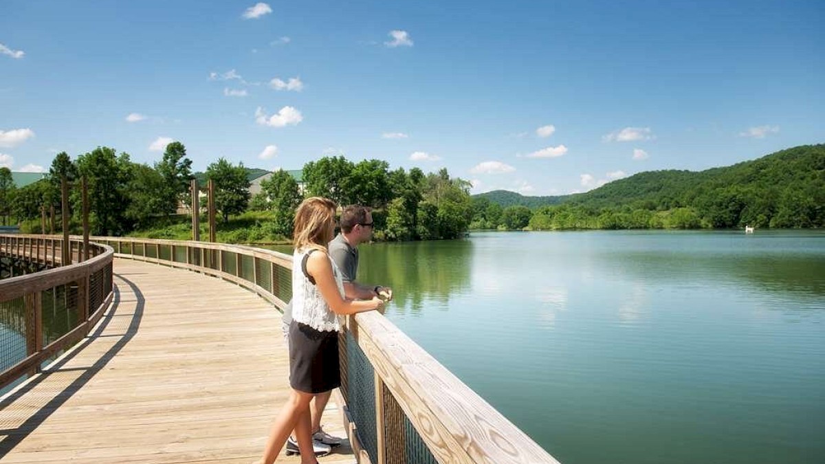 A couple stands on a wooden boardwalk overlooking a tranquil lake surrounded by lush greenery and hills under a clear, blue sky.