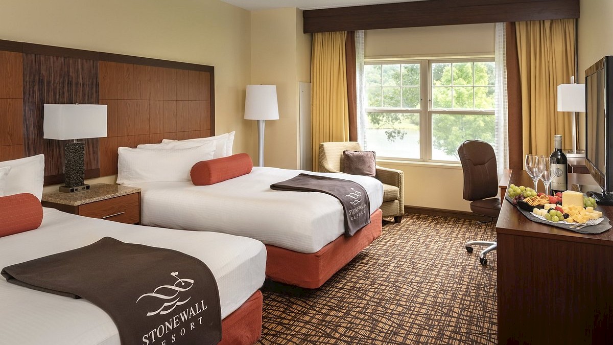 This image shows a hotel room with two beds, a window, a chair, a floor lamp, and a table with a bottle of wine and fruit.