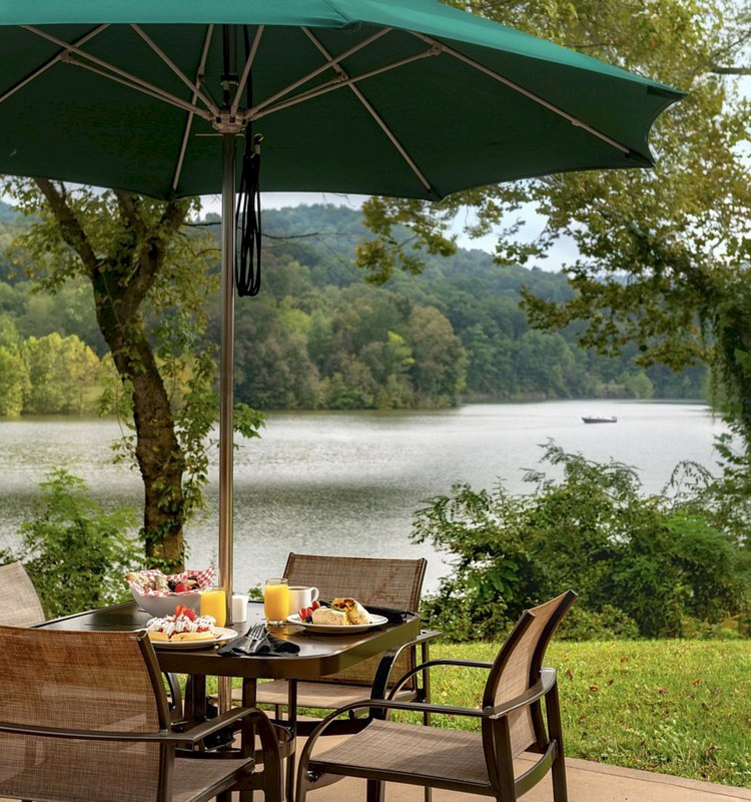 A patio table with food sits under a green umbrella, overlooking a serene lake and lush greenery, creating a peaceful outdoor dining scene.