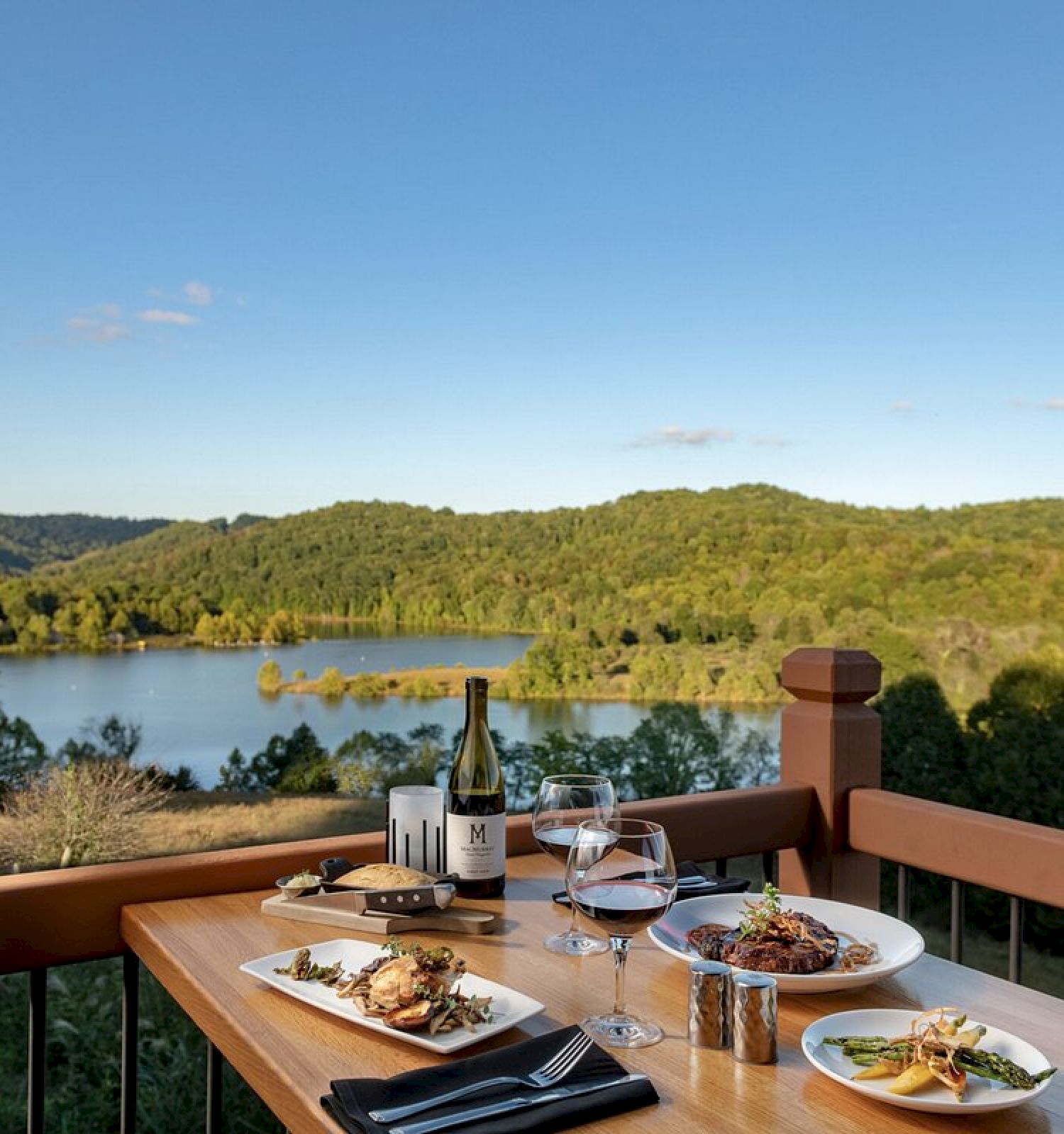 A table with food and wine overlooking a serene lake and forested hills under a clear sky, providing a peaceful dining experience.