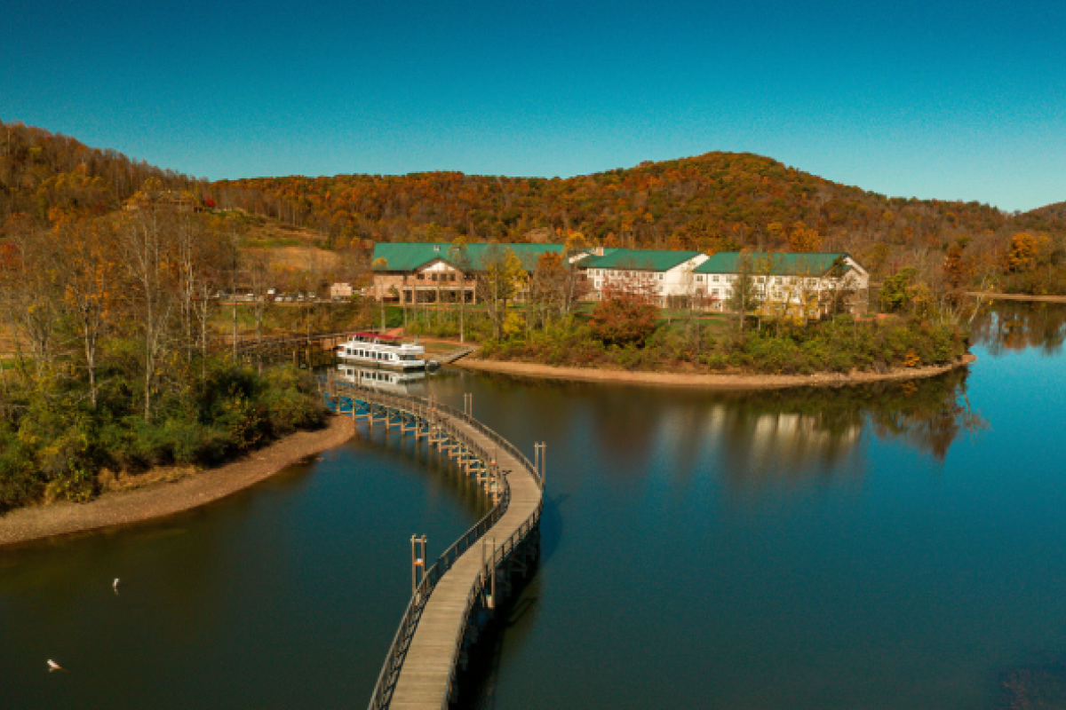 A scenic view of a lake with a curved walkway leading to buildings surrounded by autumn trees and hills under a clear blue sky.