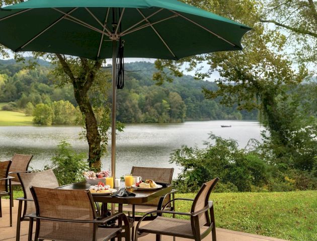 An outdoor dining setup under a green umbrella with several chairs, a table with breakfast foods, and a scenic lake view in the background.