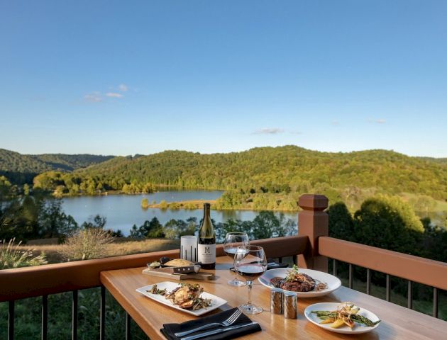 A table with plates of food, a bottle of wine, and glasses set against a scenic view of a lake and lush green hills under a clear sky.