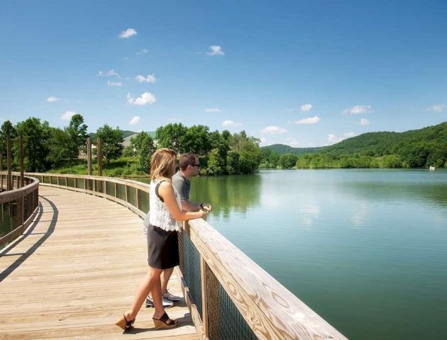 Two people stand on a wooden boardwalk overlooking a serene lake with surrounding greenery and mountains under a blue sky with scattered clouds.