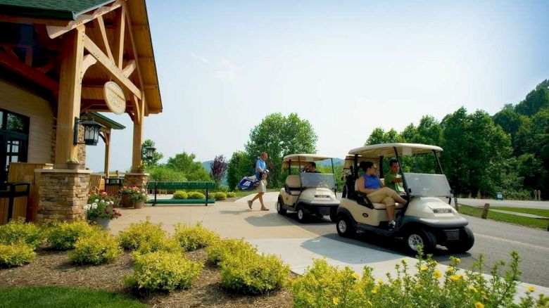 The image shows a golf course entrance with two parked golf carts and a person carrying a golf bag, surrounded by greenery and a wooden building.