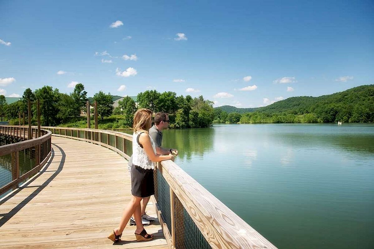 A couple stands on a wooden walkway over a serene lake, enjoying the scenic view of trees and hills under a clear, blue sky.