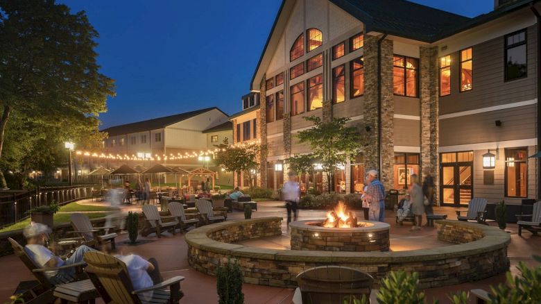 People are gathered around a cozy outdoor fire pit in the evening, with a building in the background lit up warmly.
