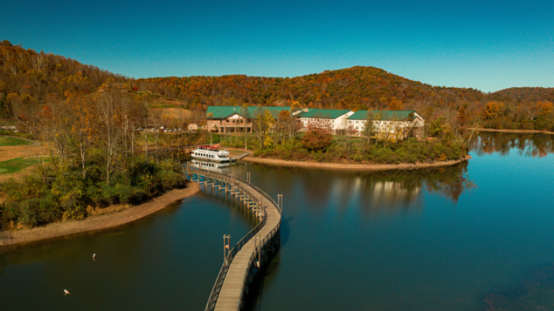 An aerial view of a scenic lake with a curved wooden pedestrian bridge leading to buildings. The surrounding area features autumn foliage and hills.