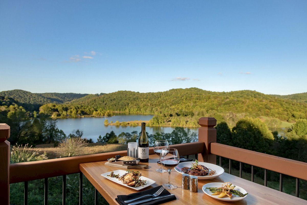 A table set with a meal, including wine and various dishes, overlooks a scenic landscape with a lake and green hills beneath a clear blue sky.