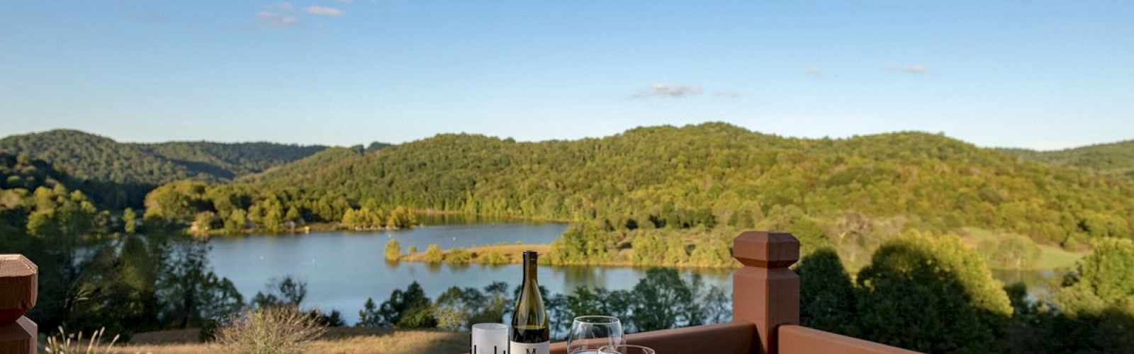 A wooden table with plates of food, wine, glasses, and cutlery overlooks a scenic lake and hilly landscape under a clear blue sky.