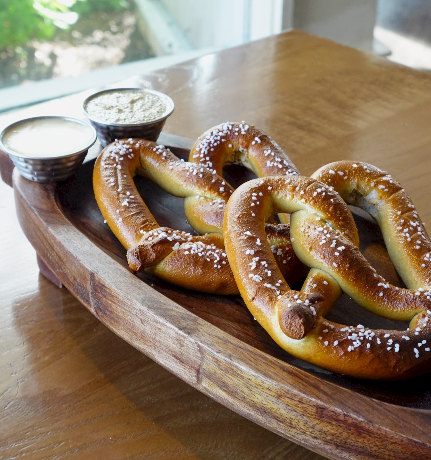 Three salted soft pretzels on a wooden tray with two small cups of dipping sauces on the side.