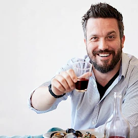 A bearded man toasting with a glass of dark drink at a table with food, smiling warmly.