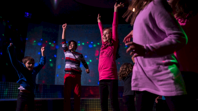 Children are dancing joyfully in a room with colorful lights, some with arms raised, wearing casual clothes.